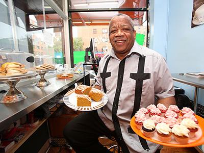 Rolando carries two trays filled with cupcakes inside of his small bakery. Rolando carries two trays filled with cupcakes inside of his small bakery.